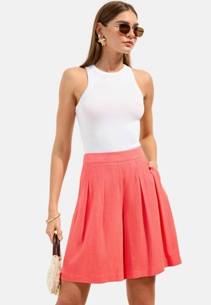 Frau trägt weißes ärmelloses Oberteil, korallenfarbenen plissierten Rock, große Blumenohrringe, braune Sonnenbrille und hält eine kleine Handtasche mit Holzgriffen.