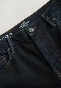 Mørkeblå denim-jeans med orange syninger, metalknap med teksten "Premium Denim Goods Since 1982" og indvendigt mærke, der angiver "Next Performance Relaxed Fit."