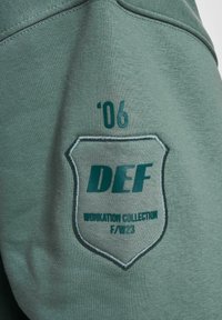 Le détail du sweat-shirt vert présente un texte brodé 'DEF', l'année '06' et 'COLLECTION WORKATION' dans un design de blason. Tissu doux et texturé.