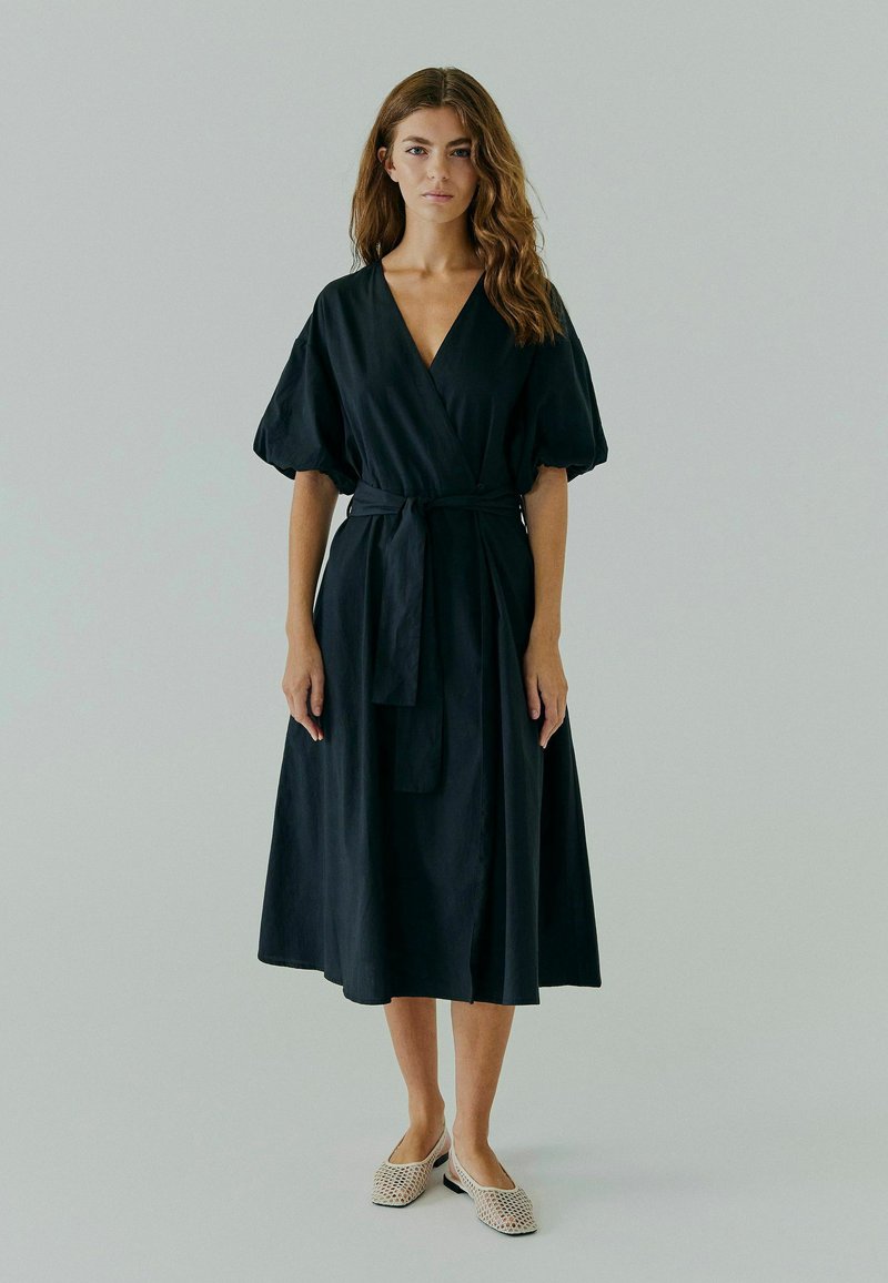 Robe noire portefeuille avec un décolleté en V plongeant, des manches bouffantes et une taille à lacer. Fabriquée en un tissu doux et léger avec un design fluide arrivant aux chevilles.
