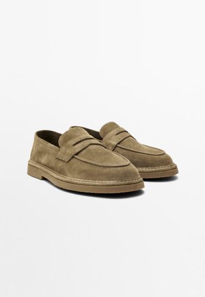 Par brune ruskind slip-on loafers med runde tæer, lave hæle og en vandret stropdetalje henover toppen, placeret mod en ensfarvet baggrund.