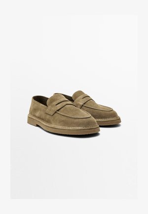 Par brune ruskind slip-on loafers med runde tæer, lave hæle og en vandret stropdetalje henover toppen, placeret mod en ensfarvet baggrund.