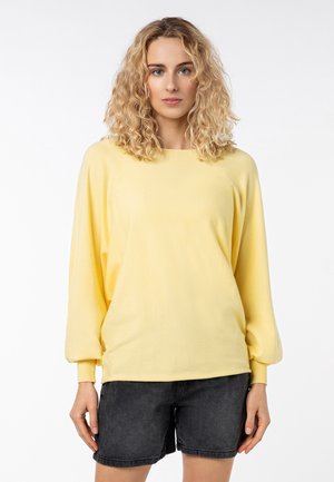 Gelber, langärmeliger Pullover mit lockerer Passform und gerippten Bündchen, kombiniert mit schwarzen Jeansshorts. Weiche Textur; einfaches, minimales Design.