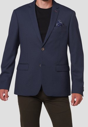 Mand iført en mørkeblå tekstureret blazer med to knapper, en sort skjorte under, mørke bukser og et blåmønstret lommetørklæde.