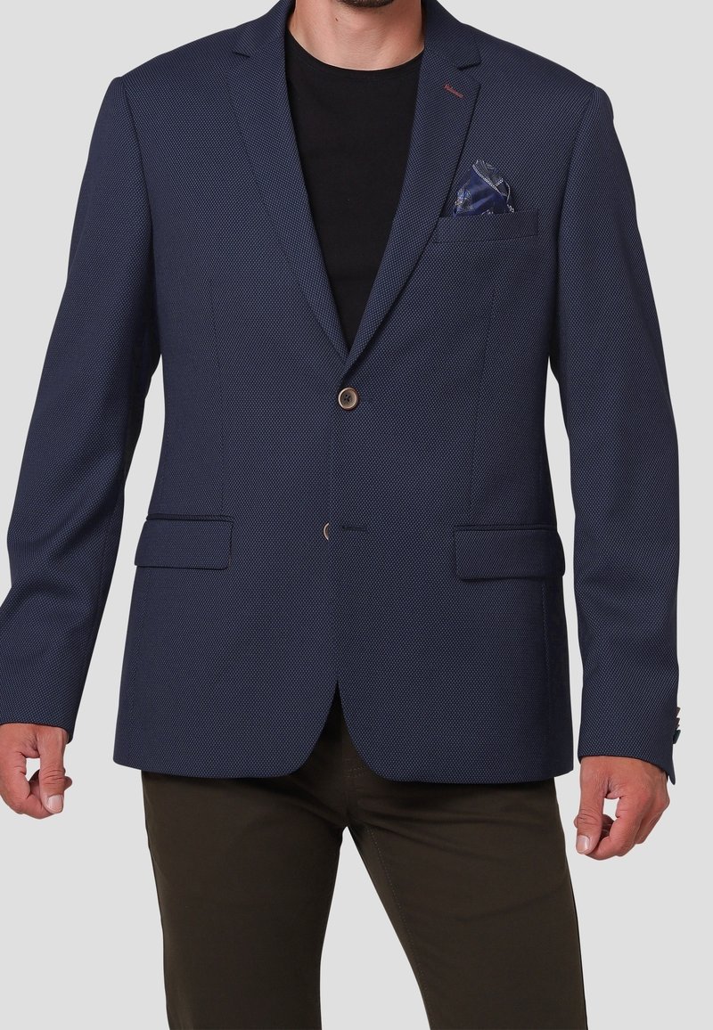 Homme portant un blazer texturé bleu foncé à deux boutons, une chemise noire en dessous, un pantalon foncé et un mouchoir de poche bleu à motif.
