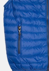 Blå quiltad jacksektion med vertikal zipficka och synliga dragkedjedetaljer, som visar tygstruktur och sömmar.