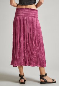 Plisovaná maxi sukně z jasně růžové látky s nařaseným pasem a hladkou texturou. Doplňena černými sandály na ploché podrážce se zapínacími pásky.