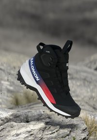 Ghete de drumeție Salomon negre și roșii cu talpă rugged, suspendate în aer deasupra unui teren stâncos, având bucle de prindere și sistem de șireturi rapide.