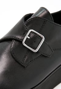 Chaussure en cuir noir avec une boucle argentée, texture lisse, forme arrondie et accents cousus le long des bords.