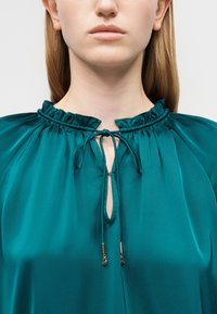 Κοντινό πλάνο γυναίκας που φορεί μπλούζα από σατέν τιρκουάζ με γιακά με βολάν και κορδόνια δεμένα στο λαιμό με χρυσά άκρα, παρουσιάζοντας το λαιμό και τους ώμους.