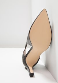 Chaussure à talon haut en cuir noir avec un bout pointu, dotée d'une sangle de cheville et d'une semelle beige, reposant sur une surface blanche.