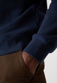 Navy blue sweatshirt with ribbed cuffs, made of soft fabric. Brown pants with a pocket are partially visible, showing a hand resting inside.