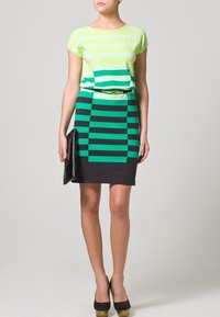 Top in maglia a righe verdi con maniche corte abbinato a una gonna a righe verdi e nere. Una pochette nera e scarpe con tacco alte marroni completano il look.