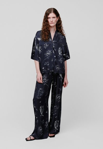 Femme aux cheveux bouclés portant une chemise ample bleu marine foncé imprimée de fleurs avec des boutons et un pantalon large assorti avec des sandales noires.