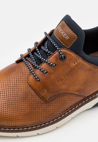 Chaussure en cuir perforé marron avec des accents bleu marine, lacets bleu et blanc, et une étiquette en cuir beige affichant "RIEKER". Semelle blanche résistante.