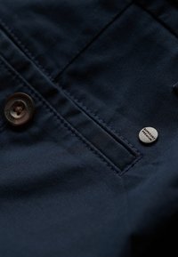 Marineblåt stof, glat tekstur, med en stor brun knap og en lille metallisk detalje med teksten "Superdry Premium."