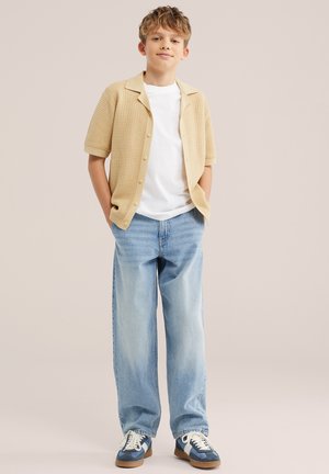 Jeune garçon debout les mains dans les poches, portant un cardigan beige en maille à manches courtes, un T-shirt blanc, un jean bleu clair et des baskets bleues.