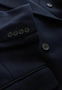 Marineblauer Wollmantel mit Schößchenkragen, vier schwarzen Knöpfen an den Ärmeln und weicher Textur mit leichtem Glanz.