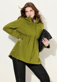 Giacca softshell verde oliva con cappuccio, vita cinchata e tasche laterali con zip. Abbinata a pantaloni neri e una clutch nera.