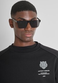 Felpa nera con collo alto, caratterizzata da una grafica di una tigre bianca e testo del marchio. Gli occhiali da sole rettangolari neri opachi completano il look.