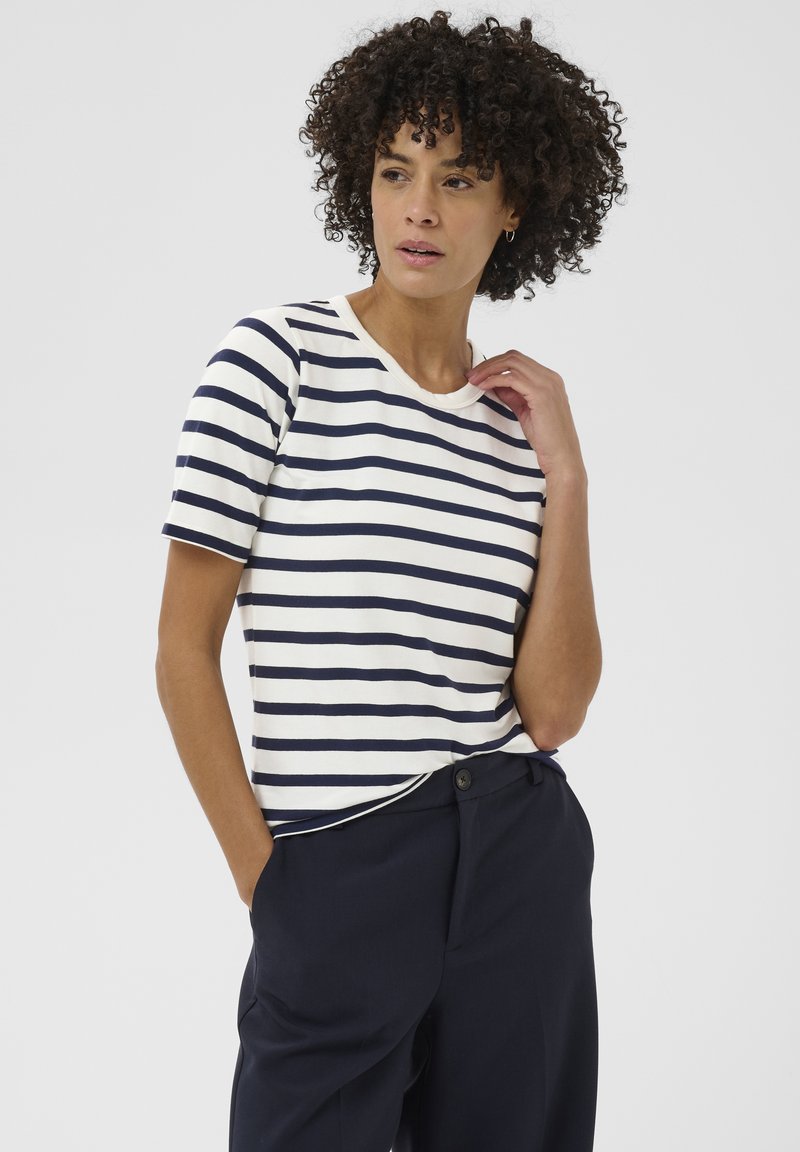 Femme aux cheveux bouclés portant un t-shirt à manches courtes rayé blanc et bleu marine et un pantalon bleu marine, une main dans la poche et l’autre près du col.