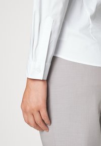 Biała koszula z długim rękawem z wyspecjalizowanym mankietem zakończonym guzikiem, zestawiona z jasnoszarymi, strukturalnymi spodniami. Materiał wydaje się gładki i lekki.
