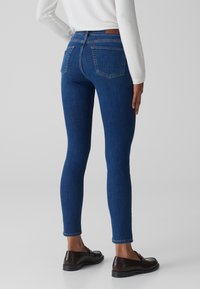 Jeans in denim blu sottili con vita media, tasche posteriori e silhouette aderente. Abbinati a mocassini neri lucidi.