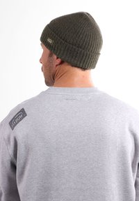 Bonnet vert en tricot côtelé avec un revers plié. Porté avec un sweat-shirt gris arborant une étiquette sur l'épaule gauche.