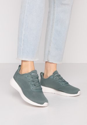 Jean bleu clair associé à des baskets Skechers grises à lacets avec semelles blanches sur une surface lisse.
