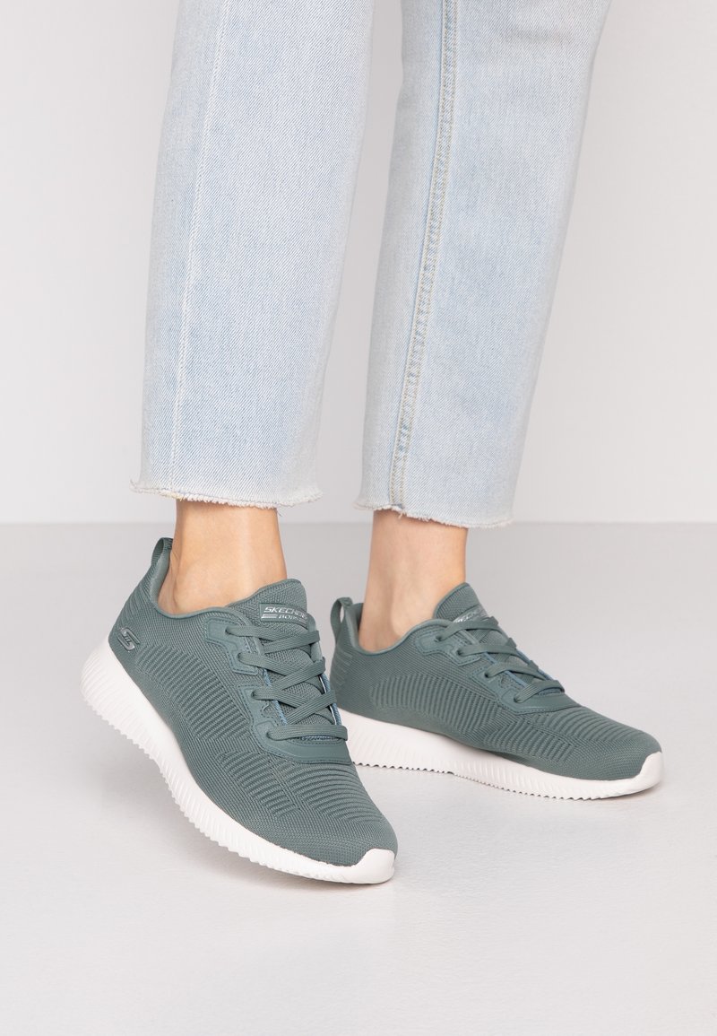 Jean bleu clair associé à des baskets Skechers grises à lacets avec semelles blanches sur une surface lisse.