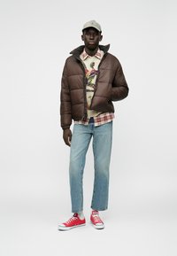 Brązowa puchowa kurtka na zamek, wzorzysty sweter pod spodem, jasnoniebieskie dżinsy i czerwone trampki na modelu na jednolitym tle.