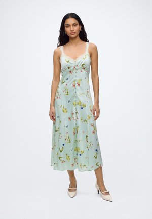 Femme aux longs cheveux foncés portant une robe midi fleurie sans manches bleu clair et des talons blancs pointus, debout sur un fond blanc.