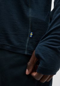 Κοντινό πλάνο σε σκούρο μακρυμάνικο πουκάμισο με ετικέτα της σουηδικής σημαίας σε μπλε και κίτρινο κοντά στη ραφή, χέρι ακουμπά στο πλάι.