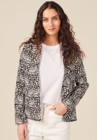 Czarna marynarka z białym wzorem liści, lekki materiał, otwarty przód, długie rękawy, noszona na białym t-shircie, w połączeniu z jasnymi spodniami.