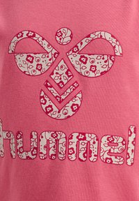 Lyserød t-shirt med et blomsterdesign af mærkenavnet "hummel" i hvidt og lyserødt bogstaver, sat på et tekstureret stof.