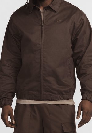 Homme portant une veste marron zippée avec des poignets élastiques et un pantalon marron assorti, debout devant un fond uni.