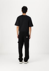 Hombre vistiendo una camiseta negra, pantalones cargo negros con una etiqueta en el bolsillo trasero y zapatillas blancas, de pie frente a una pared blanca.