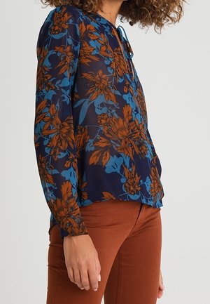 Vrouw draagt een blauwe blouse met lange mouwen met grote oranje en blauwe bloemmotieven, gecombineerd met roestkleurige aansluitende broek.