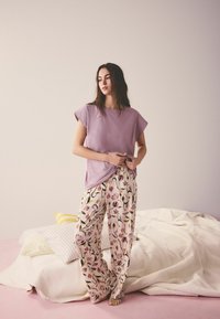 Femme debout pieds nus à côté d'un lit, portant un haut lavande ample et un pantalon large à motifs floraux dans une pièce doucement éclairée.