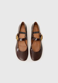 Scarpe Mary Jane in pelle verniciata marrone con punta rotonda, cinturino alla caviglia e dettaglio della fibbia dorata. Texture liscia con suola piatta e soletta imbottita.