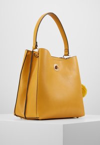 Bolso de cuero amarillo con forma estructurada, ribetes negros, herrajes dorados y un adorno de pompón. Presenta un asa superior sencilla.