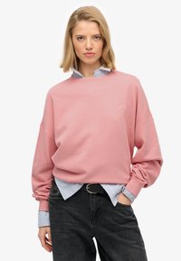 Różowy sweatshirt z okrągłym dekoltem i opadającymi ramionami, w zestawieniu z niebieską koszulą w paski. Czarne dżinsy i pasek dopełniają ten strój.