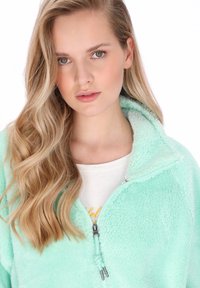 Mentaszínű polár pulóver fél cipzáras gallérral, puha tapintású, látható varrási részletekkel, fehér pólóval alatta.