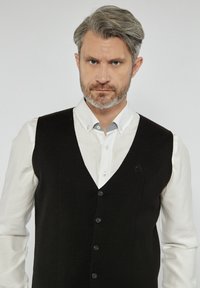 Czarna dzianinowa kamizelka z dekoltem w serek, z sześcioma guzikami i małym logo po lewej stronie, noszona na białej koszuli.