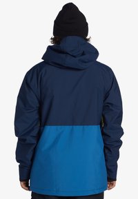 Blå- och marinblå vattentät jacka med huva, med en tvåfärgad design, elastiska ärmslut och zip-fickor på sidorna. Matt textur.