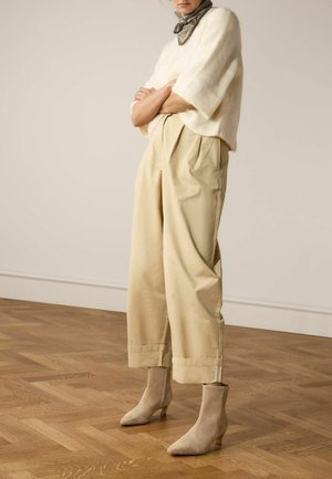 Femme debout, les bras croisés, portant un pull crème à manches courtes, un pantalon large beige raccourci, des bottines marron clair et un foulard à motif autour du cou.