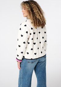 Vrouw met krullend haar in een witte gewatteerde jas met zwarte hartpatronen en blauwe jeans, die zijwaarts staat tegen een witte achtergrond.