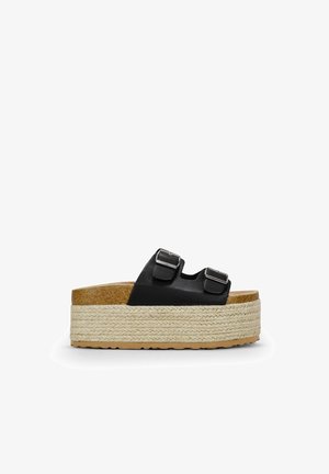 Sandales à glissière en cuir noir avec deux boucles réglables, une épaisse semelle en jute tressé, et une semelle intérieure en liège. La plateforme ajoute de la hauteur et de la stabilité.