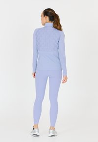 Ljust lila träningsoverdel med texturerad baksida och figurnära långärmar, matchad med leggings och silvriga träningssneakers.