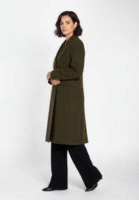 Olivgrön kappa med en texturerad yta, enkelknapp, figursydd passform och sidofickor, kombinerad med svarta vida byxor och spetsiga skor.